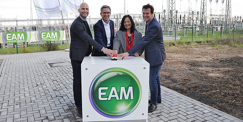 Vier Personen drücken gemeinsam einen roten Knopf auf einem weißen Pult mit dem Logo EAM im Hintergrund.