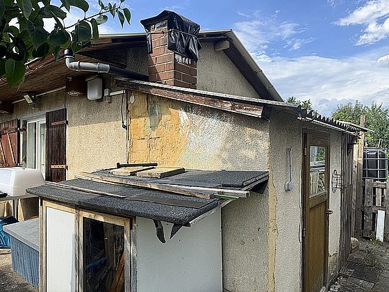 Ein einfaches, behelfsmäßig gebautes Gartenhaus mit einem Schornstein und mehreren Holzpaneelen.