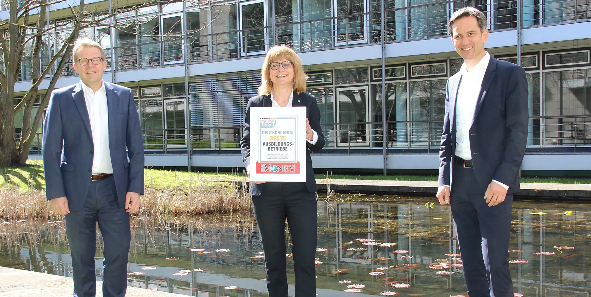 Drei Personen stehen vor einem Gebäude und halten ein Schild mit der Aufschrift Deutschlands beste Ausbildungsbetriebe in der Hand.
