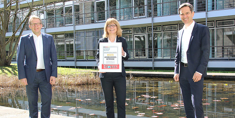 Drei Personen stehen vor einem Gebäude und halten ein Schild mit der Aufschrift Deutschlands beste Ausbildungsbetriebe in der Hand.