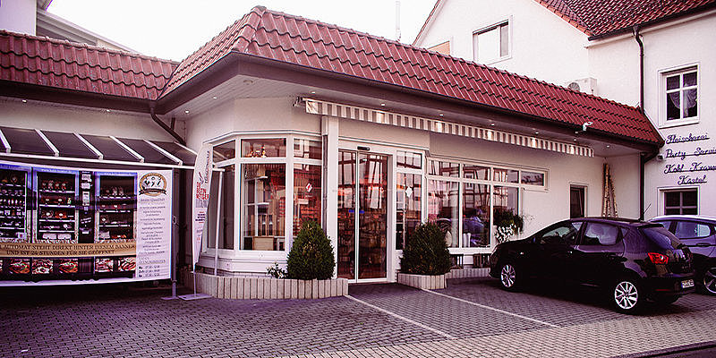 Eingang zu einem Geschäft mit großen Fenstern, begrünten Bereichen und parkenden Autos auf einem gepflasterten Platz.