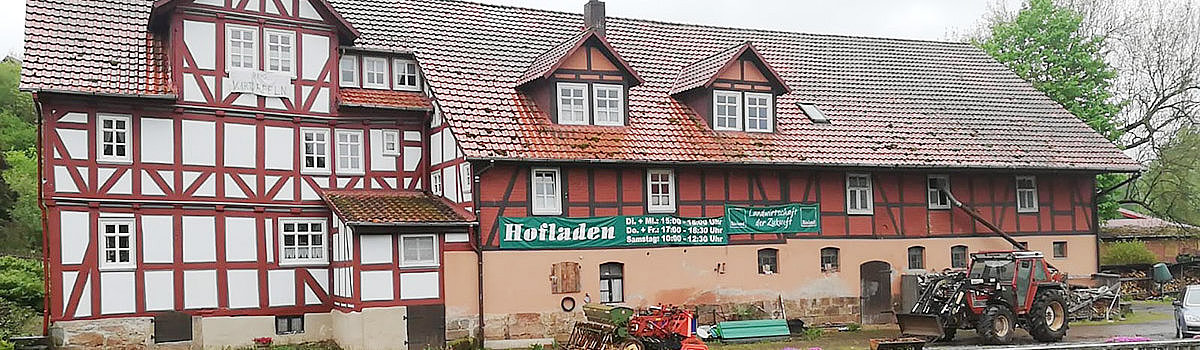 Historisches Gebäude mit Fachwerkarchitektur und einem Hofladen, umgeben von grüner Wiese und landwirtschaftlichen Fahrzeugen.