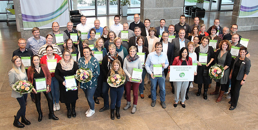 Gruppenfoto in einem modernen Gebäude, alle halten Urkunden und Blumenarrangements, mit einem Banner im Hintergrund.