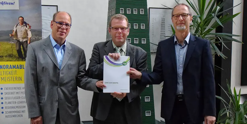 Drei Personen in Anzügen stehen zusammen und halten ein Dokument mit dem Titel Ihre EAM-Gruppe vor einem Hintergrund mit Pflanzen.