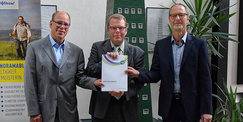 Drei Personen in Anzügen stehen zusammen und halten ein Dokument mit dem Titel Ihre EAM-Gruppe vor einem Hintergrund mit Pflanzen.
