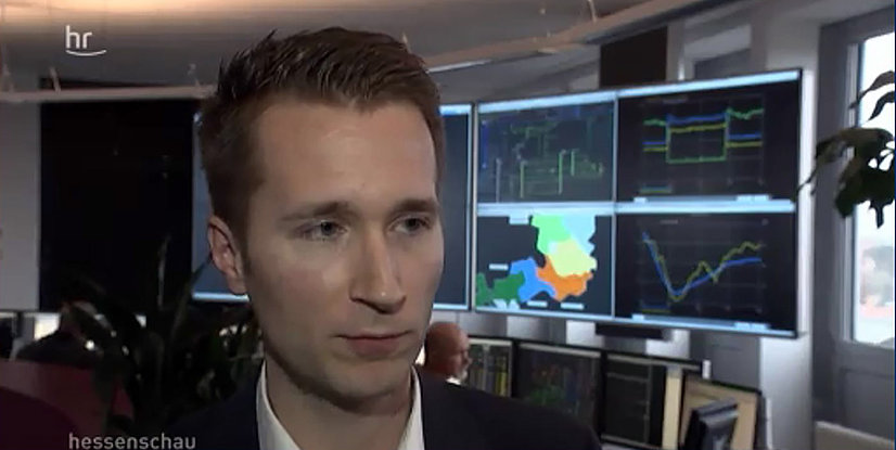 Mann mit kurzem Haar im Vordergrund, Hintergrund zeigt mehrere Bildschirme mit Grafiken und Diagrammen.
