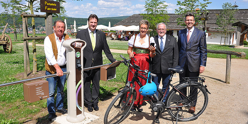 Vier Personen und ein Fahrrad stehen an einer Fahrradstation im Freien, umgeben von Bäumen und Wiese.