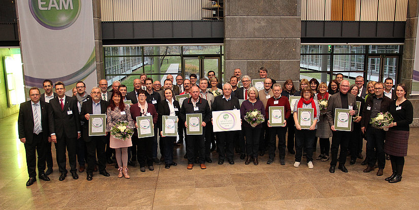 Gruppenfoto von Teilnehmern einer Preisverleihung mit Urkunden und Blumen im Foyer eines modernen Gebäudes.