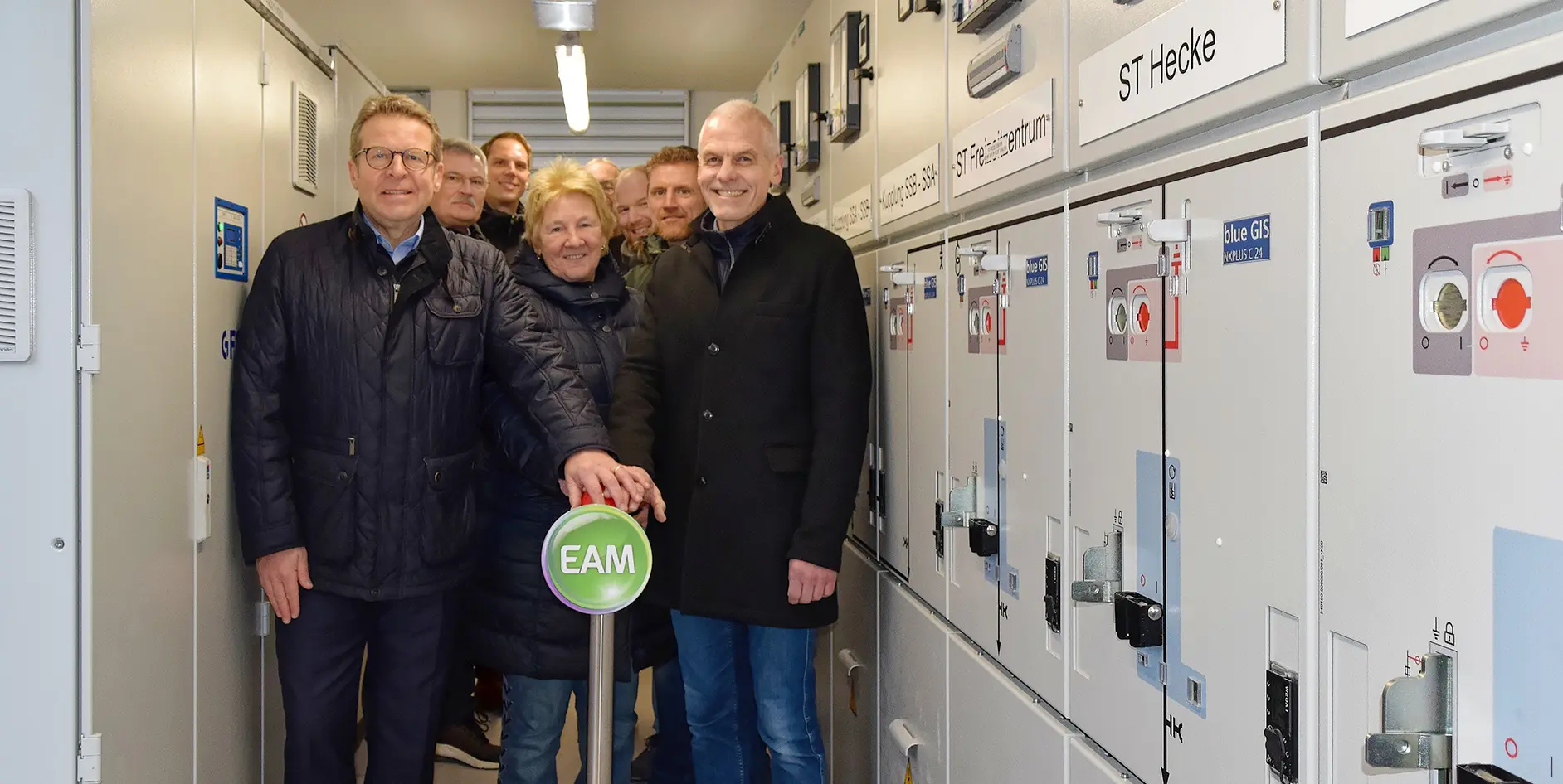 Gruppe von Personen in einem Technikraum mit Schaltschränken, die einen grünen Button mit der Aufschrift EAM betätigen.