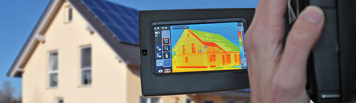 Eine Person hält ein Wärmebildgerät, das ein Haus mit Solarpanelen im Hintergrund zeigt.