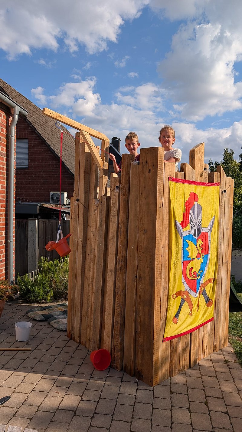 Zwei Kinder stehen auf einer Holzplattform mit einer bunten Fahne, umgeben von einem sonnigen Garten.