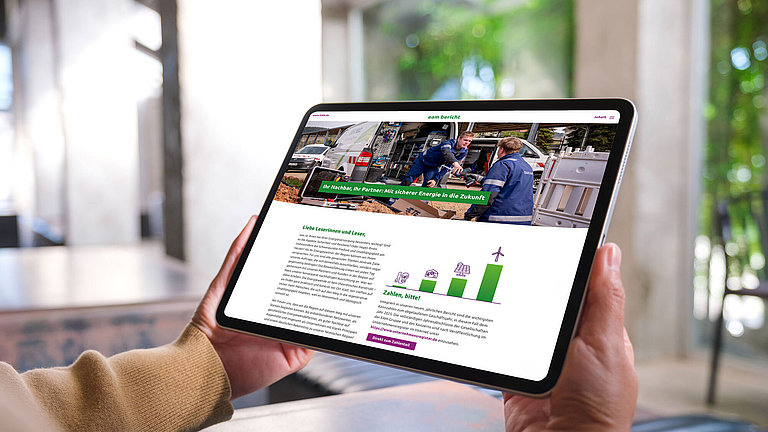Eine Person hält ein Tablet in den Händen. Auf dem Bildschirm ist die digitale Version eines EAM-Berichts zu sehen, mit einem großen Titelbild von Mitarbeitenden bei Arbeiten an einem Servicefahrzeug sowie Text- und Grafikbereichen zum Thema sichere Energieversorgung.