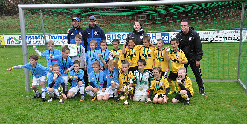 Kinderfußballmannschaft posiert stolz mit Medaillen und Pokalen vor einem Fußballtor auf einem grünen Platz.