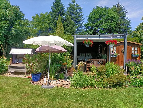 Eine grüne Gartenlandschaft mit einem Holzhaus, Gartenmöbeln, Sonnenschirmen und bunten Blumenbeeten.