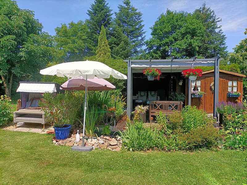 Eine grüne Gartenlandschaft mit einem Holzhaus, Gartenmöbeln, Sonnenschirmen und bunten Blumenbeeten.