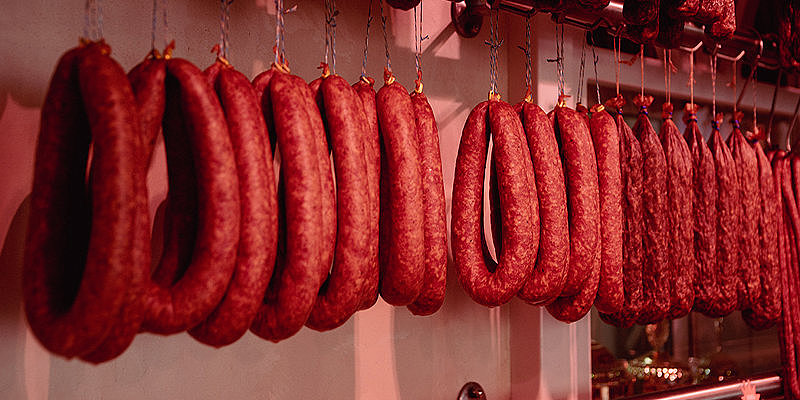Hängende Wurstwaren in verschiedenen Formen und Größen in einem Fleischgeschäft. Rotlicht betont die Farbtöne.
