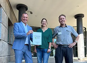 Drei Personen lächeln vor einem Gebäude und halten ein Zertifikat in der Hand.