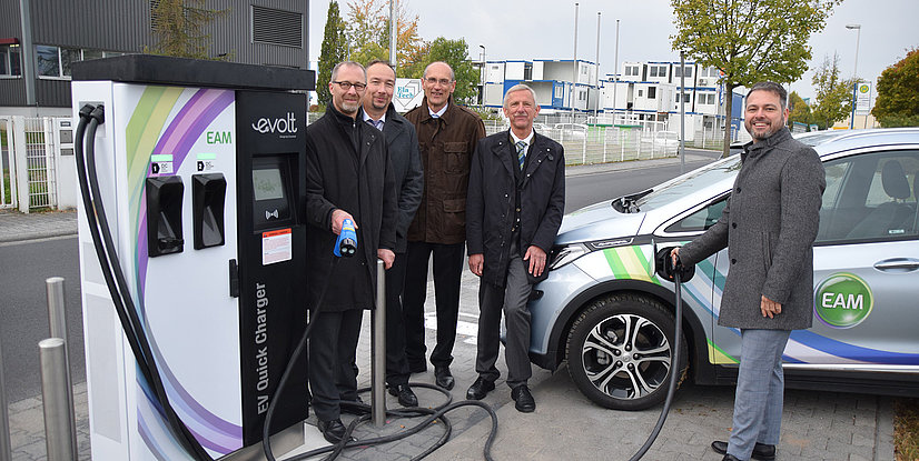 Fünf Personen stehen neben einem Elektroauto an einer Schnellladestation für Elektrofahrzeuge.
