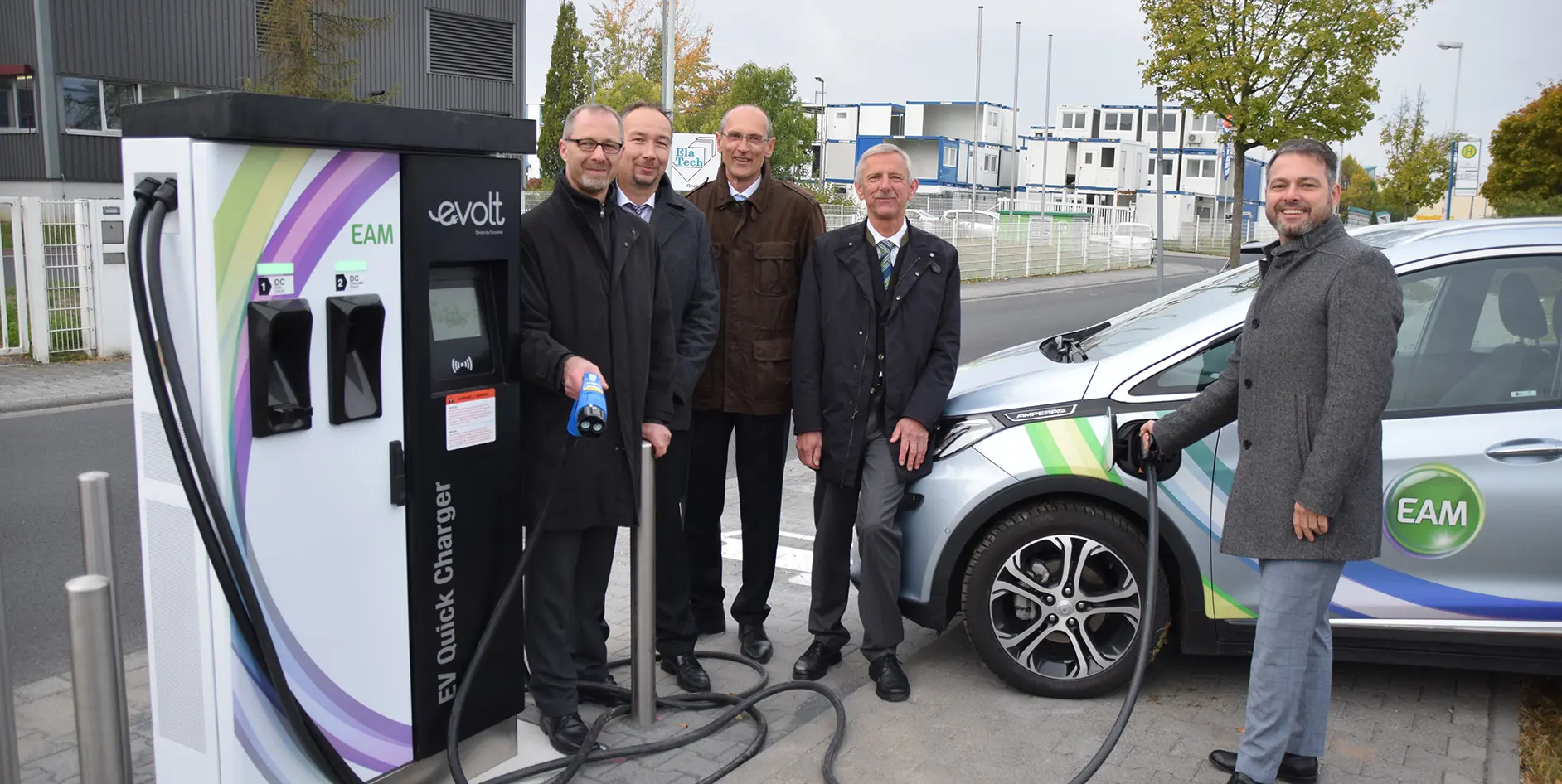 Fünf Personen stehen neben einem Elektroauto an einer Schnellladestation für Elektrofahrzeuge.