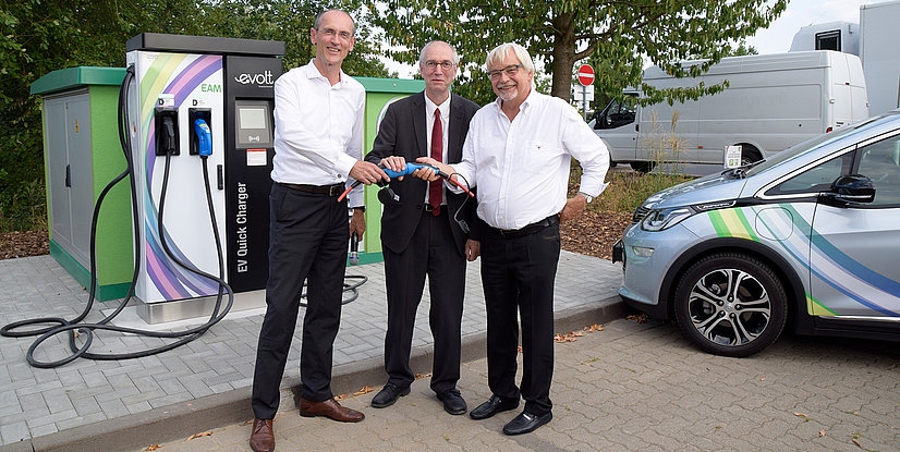Drei Personen stehen an einer Elektro-Ladestation und halten ein Ladekabel in der Hand. Im Hintergrund ist ein Elektrofahrzeug zu sehen.