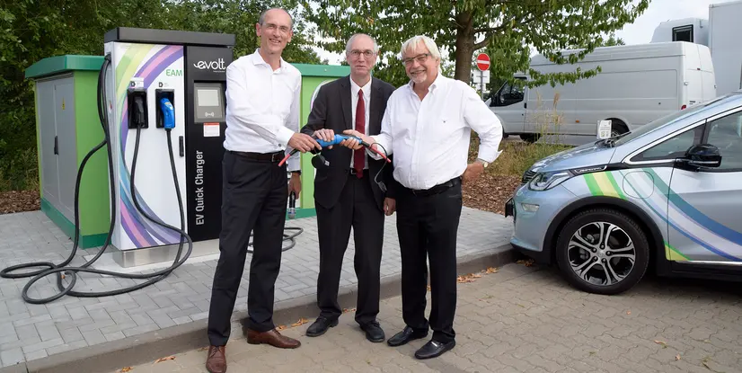 Drei Personen stehen an einer Elektro-Ladestation und halten ein Ladekabel in der Hand. Im Hintergrund ist ein Elektrofahrzeug zu sehen.