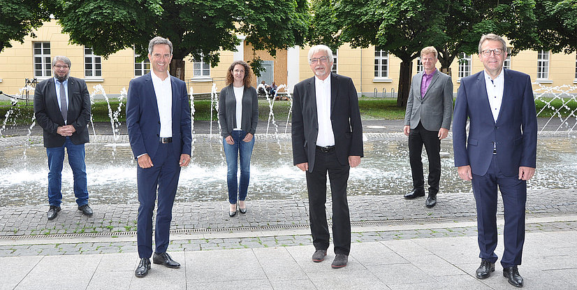 Sechs Personen stehen vor einem Brunnen in einem parkähnlichen Bereich, umgeben von Bäumen und einem Gebäude im Hintergrund.