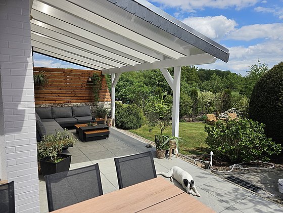 Moderne Terrasse mit Couch, Tisch, Pflanzen und Hund, umgeben von einem gepflegten Garten und blauen Himmel.