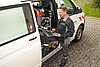Bereitschaftsdienst Techniker in Arbeitskleidung sitzt auf der Stufenleiter eines Fahrzeugs und arbeitet mit einem Laptop. Werkzeuge im Hintergrund sichtbar.