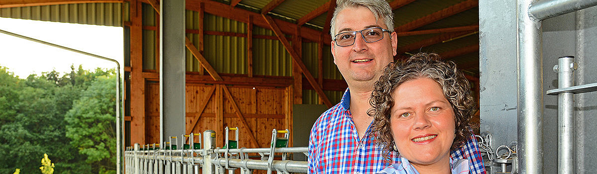 Ein Mann und eine Frau stehen in einem Stall mit Kühen im Hintergrund, umgeben von Holzwänden und natürlichem Licht.