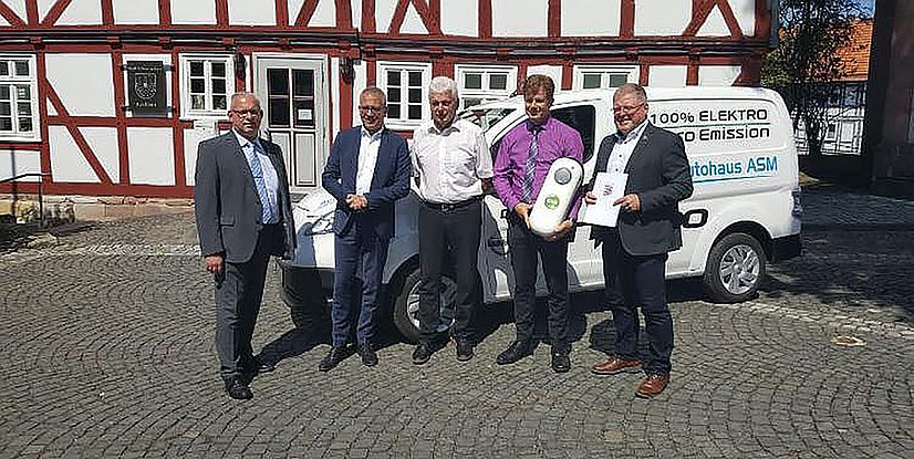 Fünf Personen stehen vor einem Elektrofahrzeug vor einem Fachwerkhaus, sie halten Dokumente und ein Objekt in der Hand.