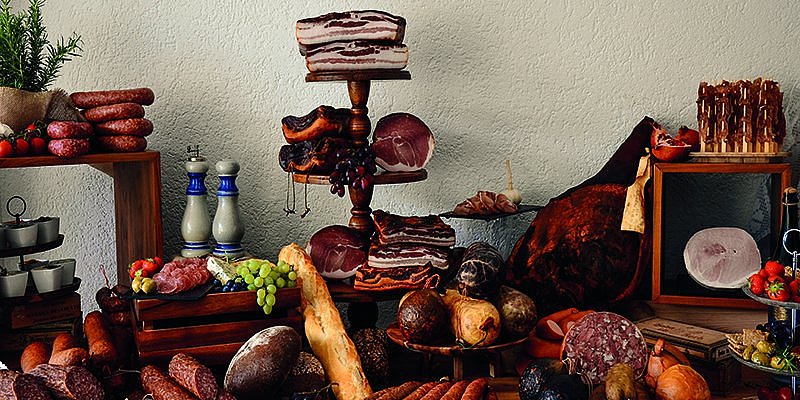 Eine Vielzahl von Wurstwaren, Schinken und frischem Brot auf einem rustikalen Holztablett, dekoriert mit Kräutern und Obst.