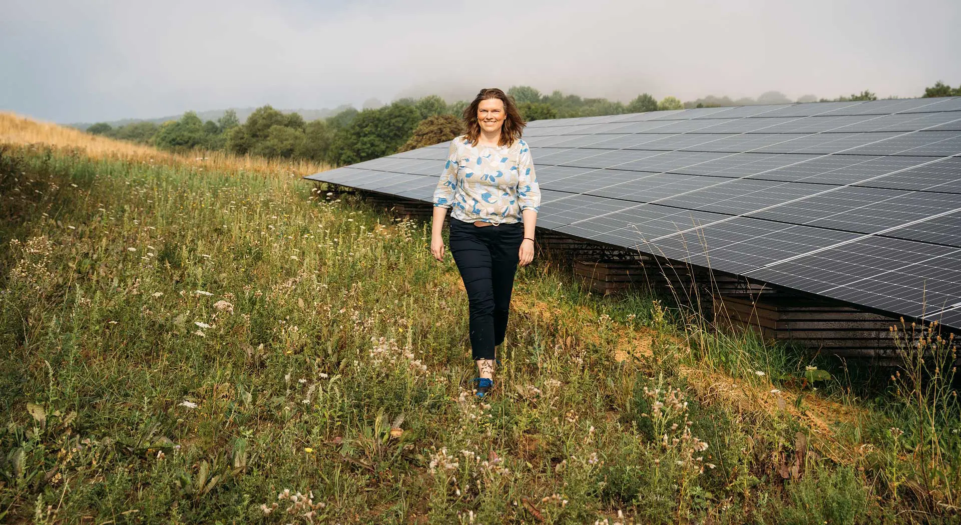  Eine lächelnde Frau steht auf einer sonnigen Wiese vor einer großen Solaranlage.