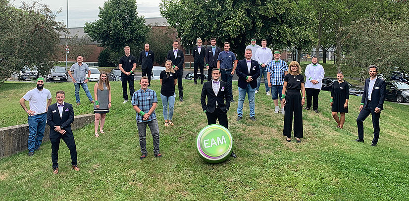 Gruppenfoto der ausgelernten Auszubildenden im Freien, verschiedene Kleidung, stehend auf einer grünen Wiese, mit einem großen EAM-Logo.