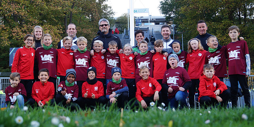 Gruppenshooting von Kindern in sportlicher Bekleidung, umgeben von Bäumen und Blumen, fröhliche Gesichter, Teamzusammenhalt.
