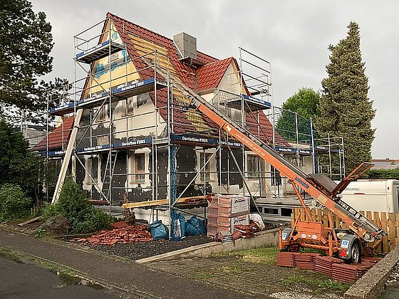 Alt-Tag: Renovierungsarbeiten an einem Einfamilienhaus mit Gerüst, Farbresten und Baustellenfahrzeug im Vordergrund.