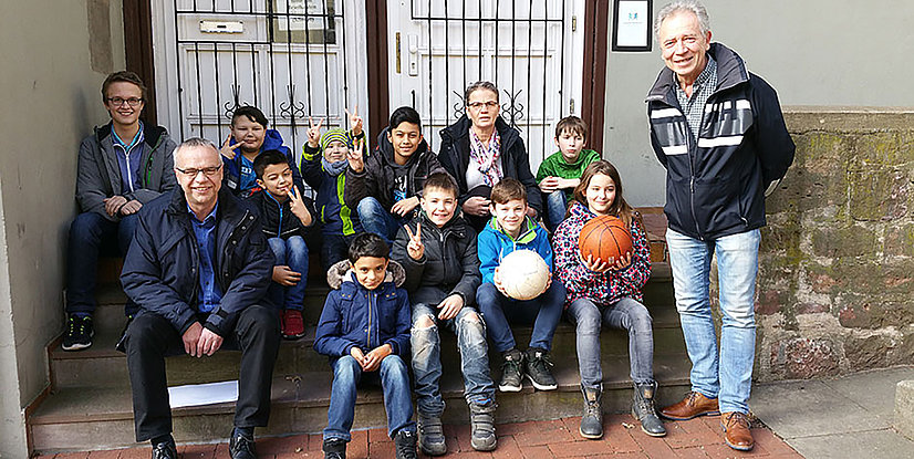 Gruppe von mehreren Kindern und Erwachsenen auf Treppen, einige halten Bälle, alle lächeln und zeigen Zeichen der Freude.
