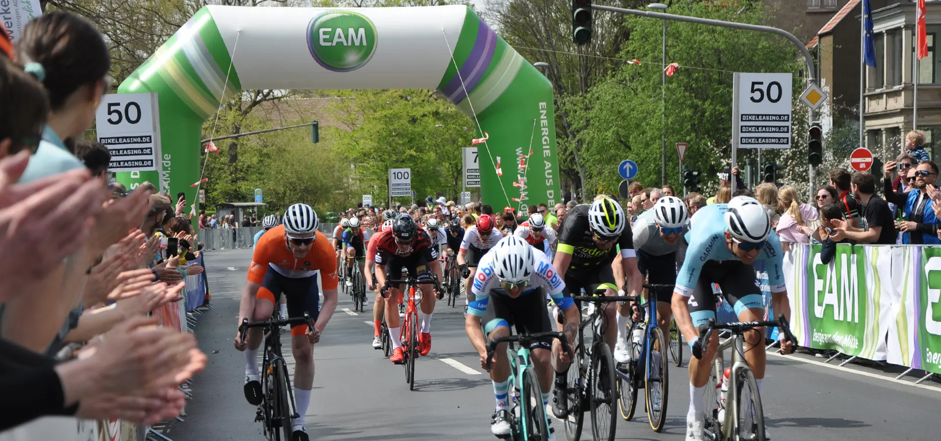 Radsportprofis beim finalen Sprint eines Rennes durch einen von der EAM gesponsorten Torbogen, im Hintergrund enthusiastische Fans die klatschen und anfeuern.