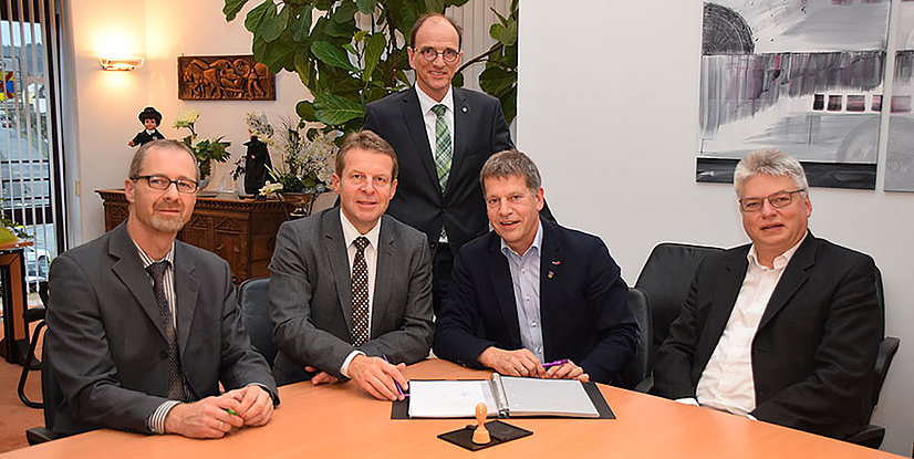 Gruppenfoto von fünf Personen in Anzügen, die an einem Tisch sitzen, mit einem Dokument und einem Stempel. Hintergrund mit Pflanzen und Kunstwerken.
