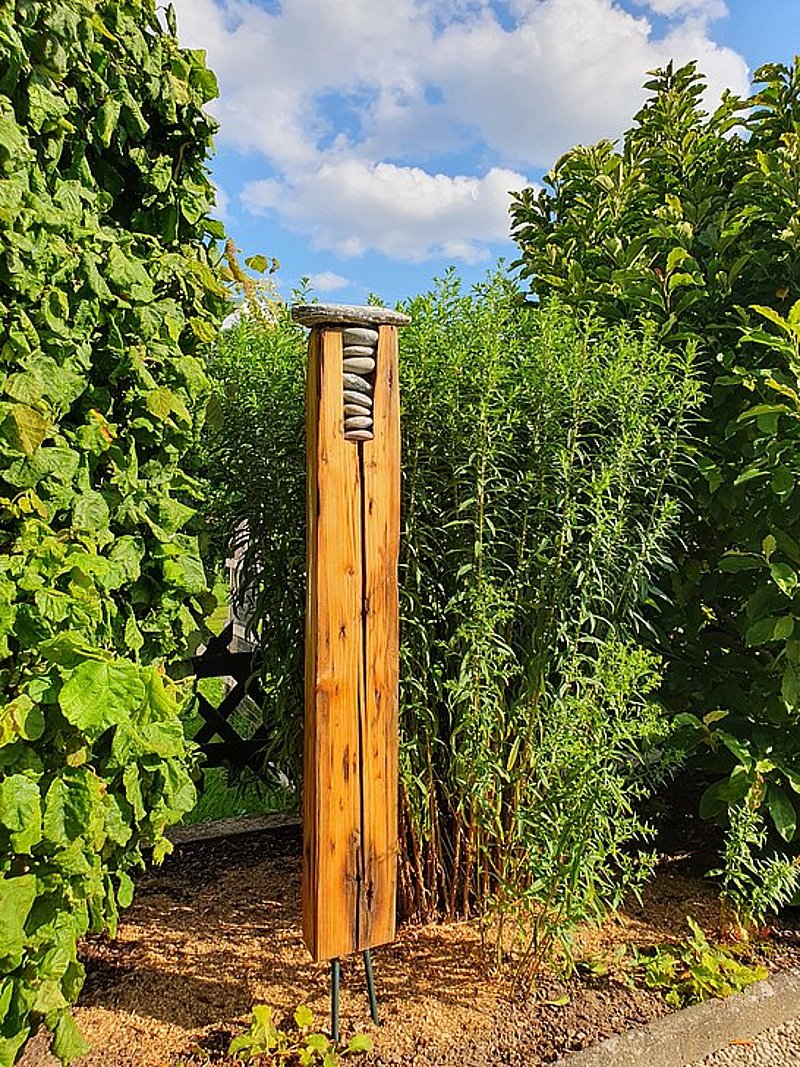 Ein Holzpfosten mit Steinen oben, umgeben von hohen grünen Pflanzen und blauem Himmel.