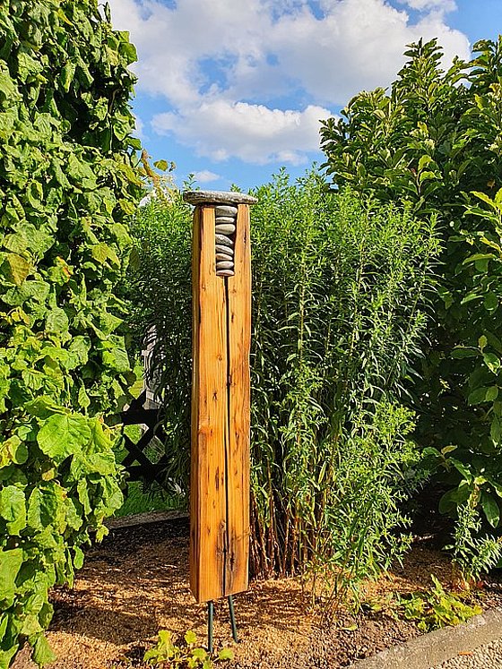 Ein Holzpfosten mit Steinen oben, umgeben von hohen grünen Pflanzen und blauem Himmel.