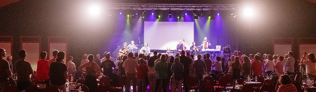 Ein Konzert mit einer Band auf der Bühne, eine Menschenmenge steht begeistert und applaudiert, Tische im Vordergrund.