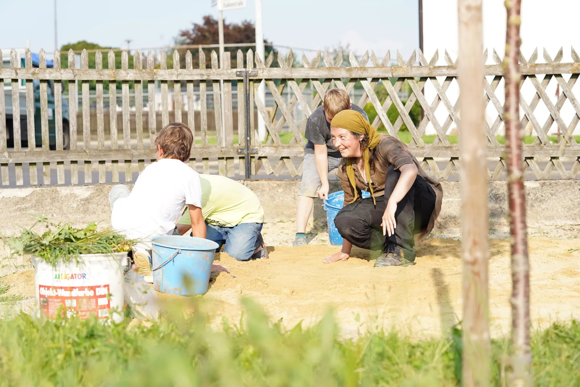 Kinder und Erwachsene arbeiten gemeinsam im Sand, umgeben von einem Zaun und Natur.