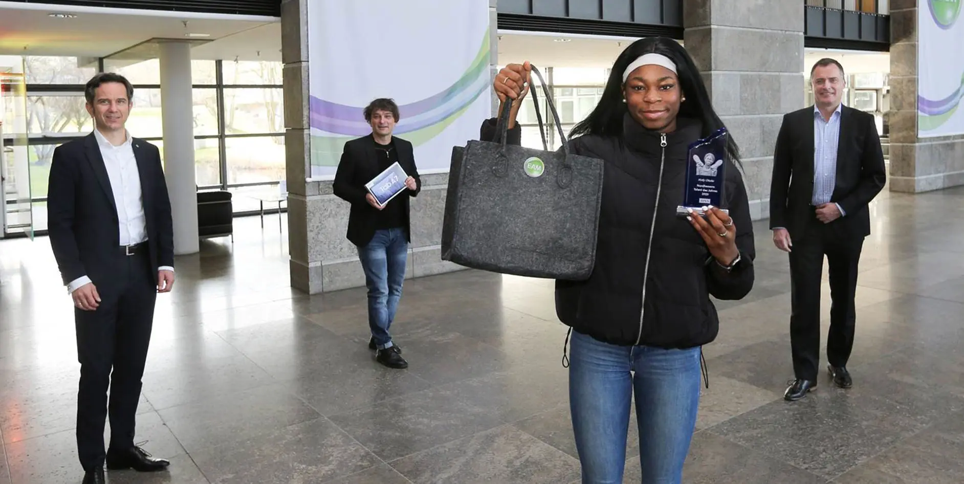  Eine junge Frau hält eine Tasche und eine Trophäe, begleitet von drei Männern die sich freuen.