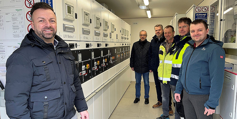 Gruppenzusammenkunft von fünf Fachleuten in einem technischen Raum mit elektrischen Geräten im Hintergrund.