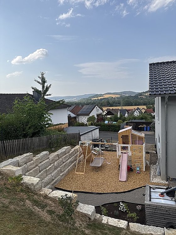 Spielplatz in einem Garten mit Schaukeln, Rutsche und Sandbox, umgeben von Wohnhäusern und Hügeln. Himmel mit wenigen Wolken.