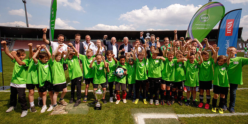 Gruppenzusammensetzung mit jubelnden Kindern in grünen Trikots, ausgestattet mit Pokalen und sportlichen Emotionen im Stadion.