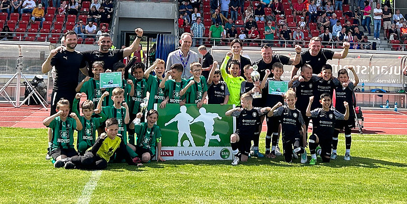 Gruppensfoto von jungen Fußballspielern mit Pokalen, mit jubelnden Coaches und einer Trophäe beim HNA-EAM-Cup.