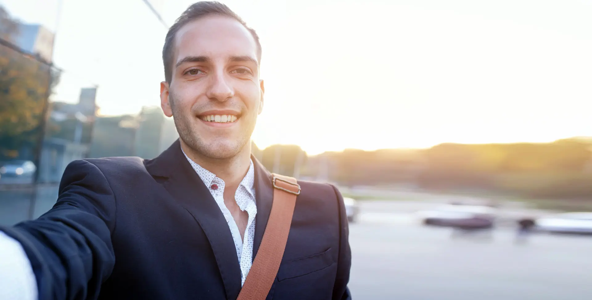 Ein lächelnder Mann im Anzug mit Umhängetasche macht ein Selfie im Freien, im Hintergrund die untergehende Sonne.