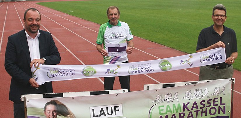 Drei Personen stehen auf einer Laufbahn, halten ein Banner des Kassel Marathons und präsentieren Medaillen.