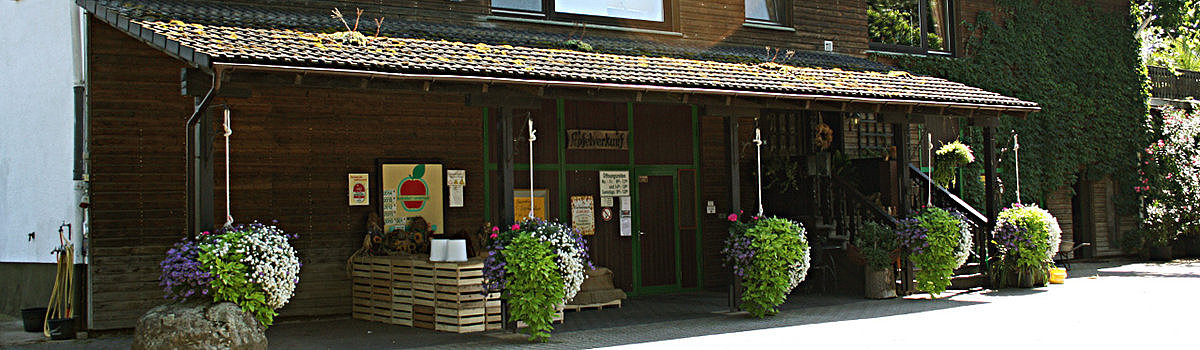 Haus mit Holzfassade, bemalten Früchten an der Wand, Blumenampeln und Eingangsbereich in sonniger Umgebung.